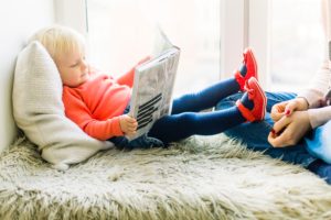 young child saying on pillow on bed with book with feet propped on moms lap smiling